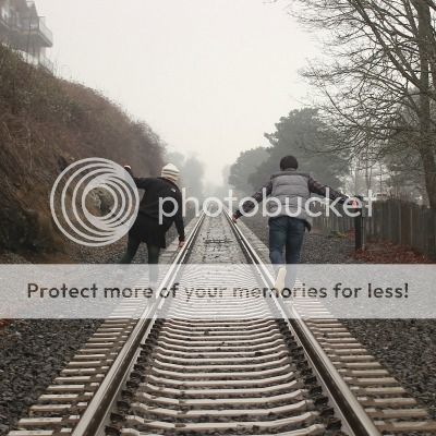 A man and a woman walking down straight railway tracks, balancing on the rails