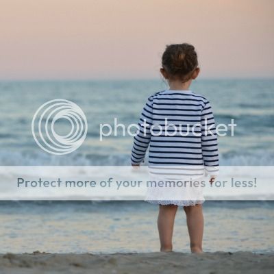 A photo og little girl standing at the water's edge on the beach, looking out to the horizon, taken from behind